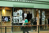 A shop window display featuring multiple white t-shirts printed with artistic black and white photographs. The storefront includes hanging t-shirts and several photographs attached to the glass. A television screen displays an image of a person in sunglasses. A person wearing a green shirt is walking past the display, holding a camera.