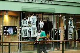 A shop window display featuring multiple white t-shirts printed with artistic black and white photographs. The storefront includes hanging t-shirts and several photographs attached to the glass. A television screen displays an image of a person in sunglasses. A person wearing a green shirt is walking past the display, holding a camera.