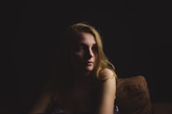 A woman with blonde hair is seated on a couch, partially illuminated by soft lighting that highlights her face while the surroundings remain in shadow. Her expression is contemplative, and the dim, focused lighting creates a sense of intimacy.