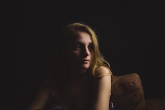 A woman with blonde hair is seated on a couch, partially illuminated by soft lighting that highlights her face while the surroundings remain in shadow. Her expression is contemplative, and the dim, focused lighting creates a sense of intimacy.