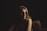 A woman with blonde hair is seated on a couch, partially illuminated by soft lighting that highlights her face while the surroundings remain in shadow. Her expression is contemplative, and the dim, focused lighting creates a sense of intimacy.