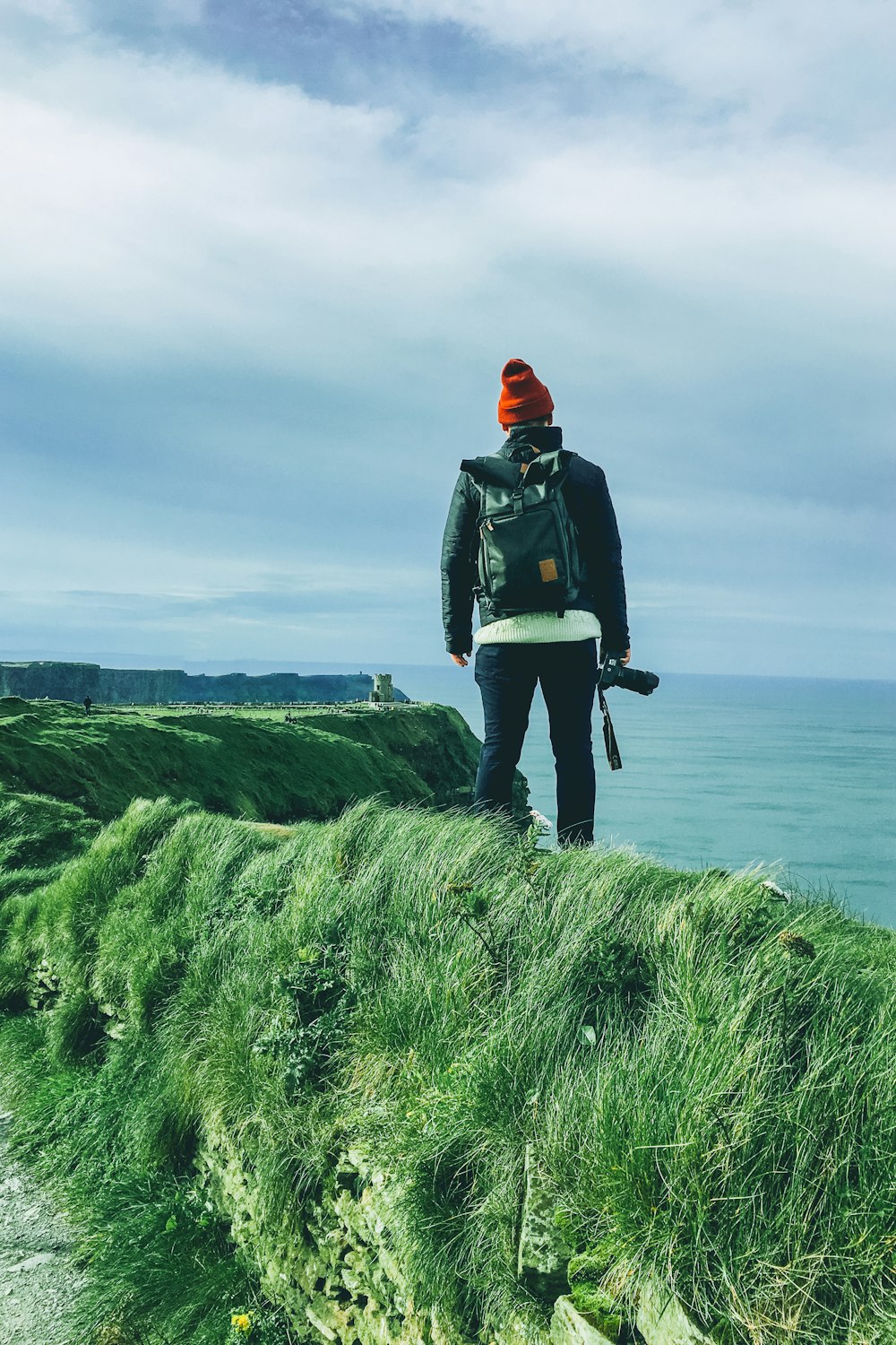 standing man holding DSLR camera on cliff looking to ocean under white sky