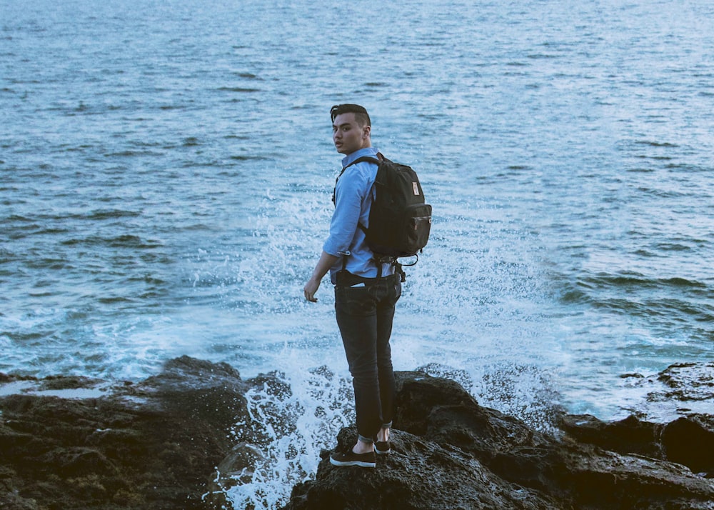 man standing on gray rock formation near body of water wearing backpack