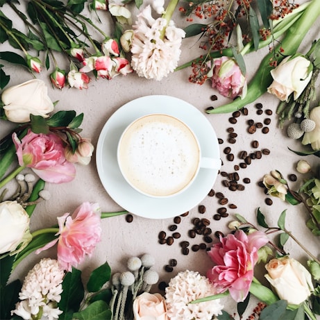A white cup filled with frothy coffee is placed in the center, surrounded by an assortment of pink and white flowers along with green leaves. Scattered coffee beans are arranged around the cup, enhancing the aesthetic arrangement. The background is a light, neutral surface.