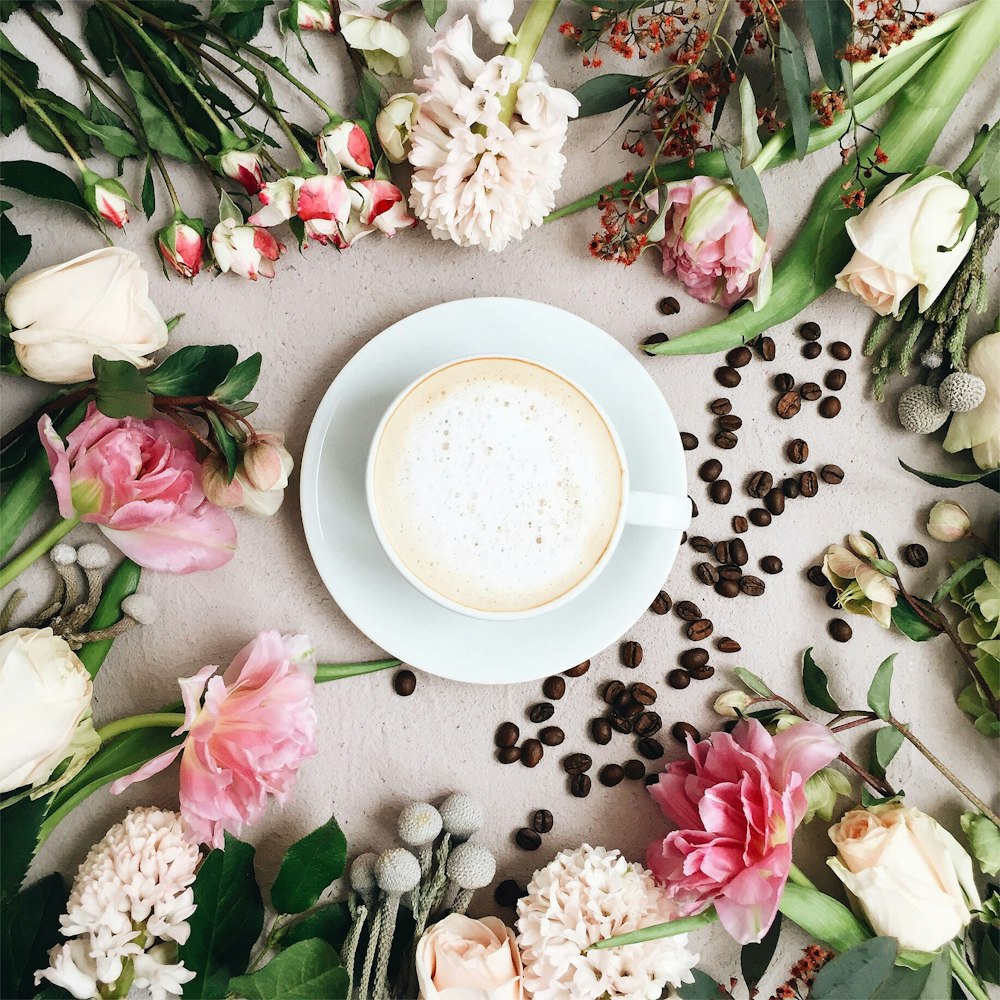 cappuccino filled white ceramic coffee cup on white ceramic saucer surrounded by assorted-type of flowers and coffee beans