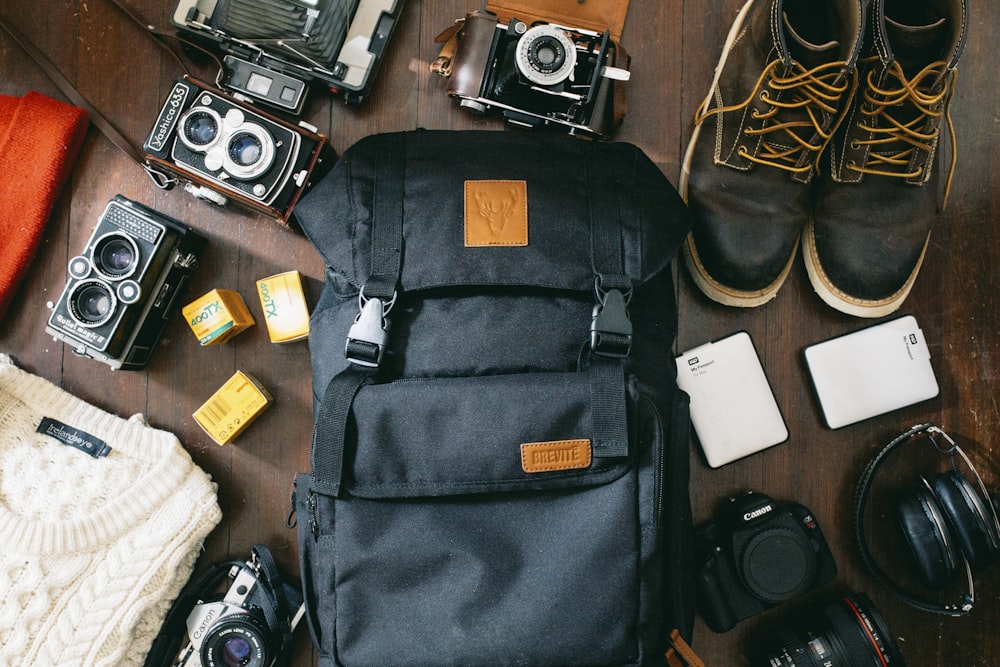 high-angle photo of black bag beside cameras, portable HDD and black leather lace-up boots