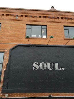 A brick building facade with large black signage displaying the word 'SOUL.' in white uppercase letters. Two windows with reflective glass are visible on the upper level. Green lamps protrude above the signage, adding a touch of industrial style.
