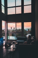 woman sitting on corner sofa beside coffee table near flat screen TV inside living room with glass window walls