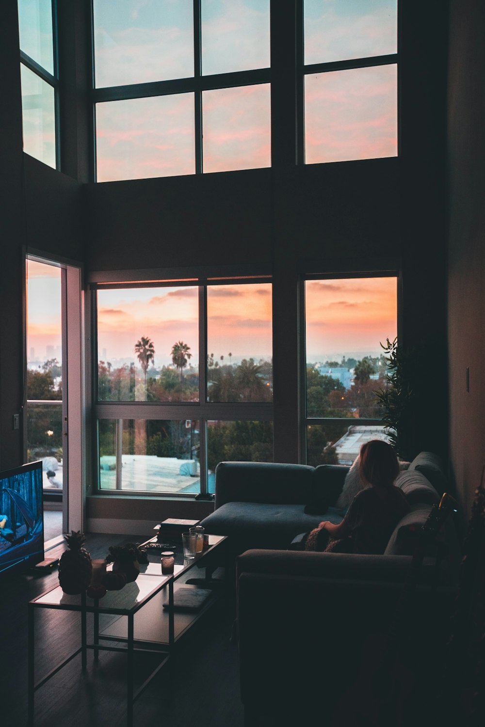 mujer sentada en el sofá de la esquina al lado de la mesa de café cerca del televisor de pantalla plana dentro de la sala de estar con las paredes de la ventana de vidrio