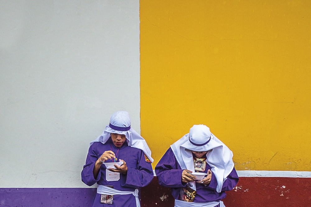 two standing men wearing purple long-sleeved top leaning on white, yellow, purple, and red painted wall