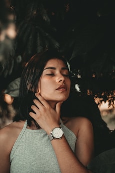 A person with closed eyes, standing amidst lush greenery. They have short, dark hair and are wearing a sleeveless, gray top. Their hand is gently resting on their neck, displaying a wristwatch. The soft lighting creates a serene and introspective atmosphere.
