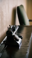 A vintage rotary telephone is placed on a sleek, dark wooden desk. The background features a light-colored textured wall, and there is a blurred green upholstered chair in the distance.
