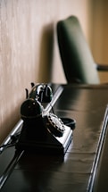 A vintage rotary telephone is placed on a sleek, dark wooden desk. The background features a light-colored textured wall, and there is a blurred green upholstered chair in the distance.