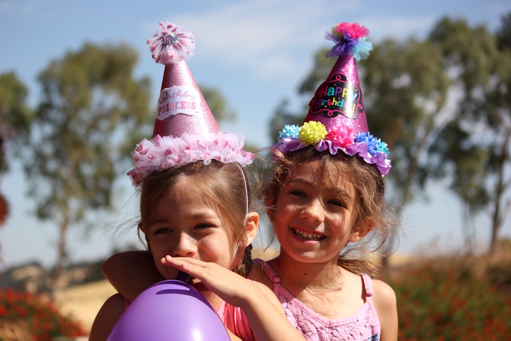 분홍색 생일 모자를 쓰고 있는 소녀 옆에 보라색 풍선을 부는 왼쪽의 소녀