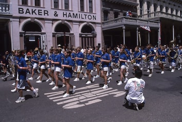 A marching band of young people dressed in blue shirts and white shorts parades down a city street. They are playing musical instruments like saxophones and clarinets as part of a procession. The street is lined with buildings and a photographer kneels in the foreground to capture the scene. Spectators are visible in the background.