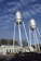 Two tall metal water towers stand on a clear day with a backdrop of blue sky and light clouds. The towers are supported by metal frameworks, and a building labeled 'Municipal Water Works' is situated beneath them, surrounded by trees and vegetation.