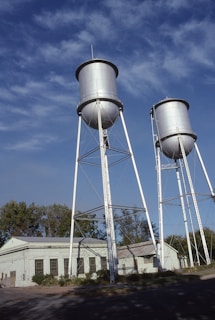 Two tall metal water towers stand on a clear day with a backdrop of blue sky and light clouds. The towers are supported by metal frameworks, and a building labeled 'Municipal Water Works' is situated beneath them, surrounded by trees and vegetation.