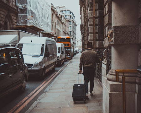 A person walks down a city street pulling a suitcase. There are several vehicles, including white vans, parked along the street, and a distinctive red double-decker bus is visible at a distance. The architecture features classical stone buildings, giving a historical feel to the scene.
