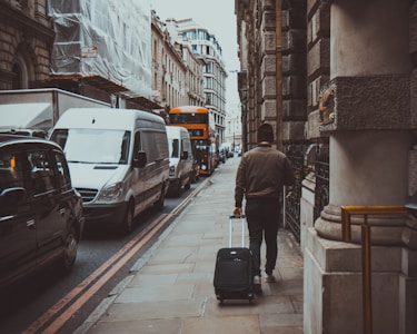 A person walks down a city street pulling a suitcase. There are several vehicles, including white vans, parked along the street, and a distinctive red double-decker bus is visible at a distance. The architecture features classical stone buildings, giving a historical feel to the scene.