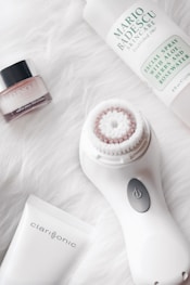 A collection of skincare and beauty products on a soft, white, fluffy background. The main items include a Clinique skin care jar, a Mario Badescu facial spray bottle, and a Clarisonic face cleansing brush with its accompanying tube. The arrangement is clean and aesthetically pleasing.