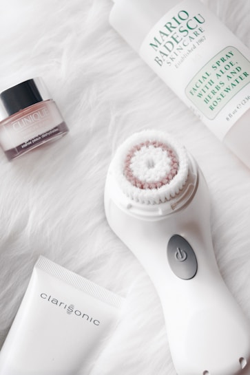 A collection of skincare and beauty products on a soft, white, fluffy background. The main items include a Clinique skin care jar, a Mario Badescu facial spray bottle, and a Clarisonic face cleansing brush with its accompanying tube. The arrangement is clean and aesthetically pleasing.