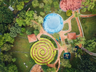 An aerial view of a well-maintained park featuring a circular garden maze. The park is lined with lush green trees and has pathways interconnecting different sections. There are two small structures with red roofs, likely gazebos. The vibrant blue circular area could be a splash pad or small water feature, surrounded by paved walkways.