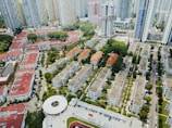 Aerial view of an urban residential area filled with mid-rise apartment buildings. These structures have a mix of red and gray roofs, surrounded by lush greenery and organized in a grid pattern. In the background, tall skyscrapers tower over the residential blocks, and a circular building can be seen in the foreground. The streets are lined with parked vehicles, and the area is interspersed with small patches of trees and grass.