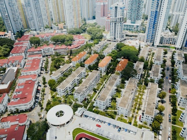 Aerial view of an urban residential area filled with mid-rise apartment buildings. These structures have a mix of red and gray roofs, surrounded by lush greenery and organized in a grid pattern. In the background, tall skyscrapers tower over the residential blocks, and a circular building can be seen in the foreground. The streets are lined with parked vehicles, and the area is interspersed with small patches of trees and grass.