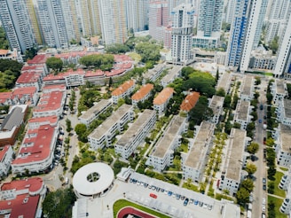 Aerial view of an urban residential area filled with mid-rise apartment buildings. These structures have a mix of red and gray roofs, surrounded by lush greenery and organized in a grid pattern. In the background, tall skyscrapers tower over the residential blocks, and a circular building can be seen in the foreground. The streets are lined with parked vehicles, and the area is interspersed with small patches of trees and grass.