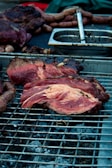 A close-up view of a barbecue grill with various types of meat, including a large piece of red meat prominently displayed in the foreground. Sausages are visible in the background, along with a metal container and tongs. The grill is situated over hot coals.