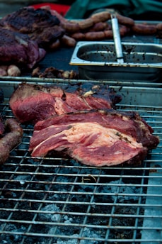 A close-up view of a barbecue grill with various types of meat, including a large piece of red meat prominently displayed in the foreground. Sausages are visible in the background, along with a metal container and tongs. The grill is situated over hot coals.
