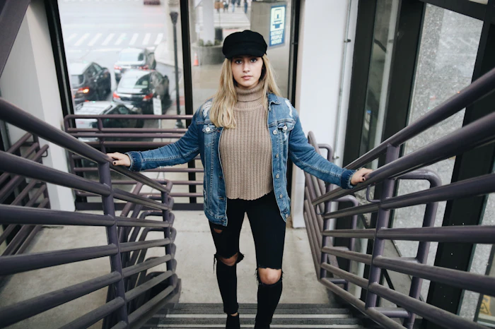Woman in a denim jacket, brown sweater and hat on stairs