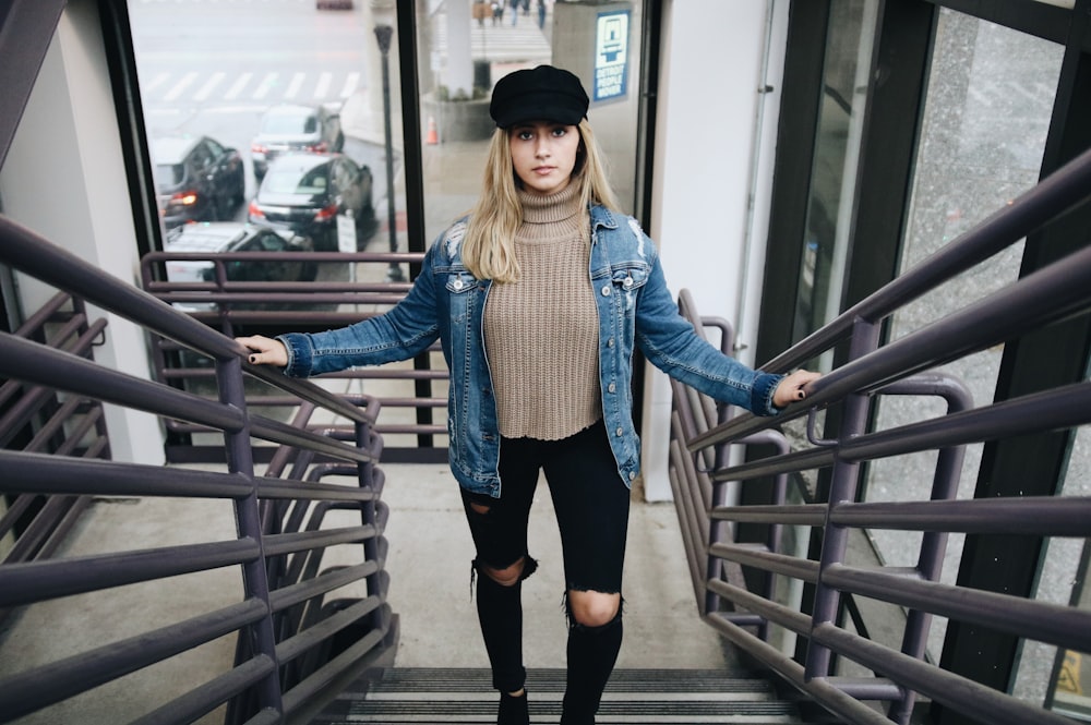 woman wearing blue denim jacket, brown sweater, black jeans, and hat standing in stair