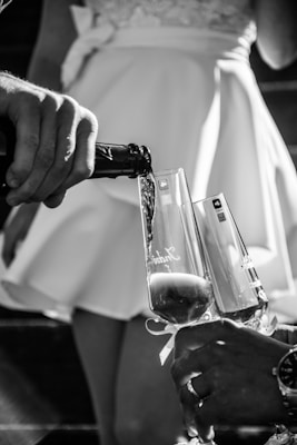 A black and white photograph captures a close-up of two champagne flutes being filled by a hand pouring from a bottle. The background features a person wearing an elegant dress, slightly out of focus. Shadows and highlights enhance the elegant and celebratory mood.