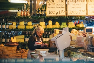 A market vendor stands behind a counter filled with various types of cheese. Shelves behind her are stocked with cheese wheels and wine bottles. She wears an apron and appears to be slicing or preparing cheese for display. The signage above displays prices and descriptions for the cheese. The market has a warm and rustic ambiance with green decorative elements.