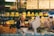 A market vendor stands behind a counter filled with various types of cheese. Shelves behind her are stocked with cheese wheels and wine bottles. She wears an apron and appears to be slicing or preparing cheese for display. The signage above displays prices and descriptions for the cheese. The market has a warm and rustic ambiance with green decorative elements.
