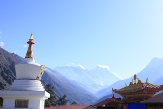 A stunning mountain landscape features snow-capped peaks in the background. In the foreground, a white Buddhist stupa with a golden spire and colorful decorations stands on the left, while a richly decorated temple roof is on the right. Trees can be seen at the base of the mountains.
