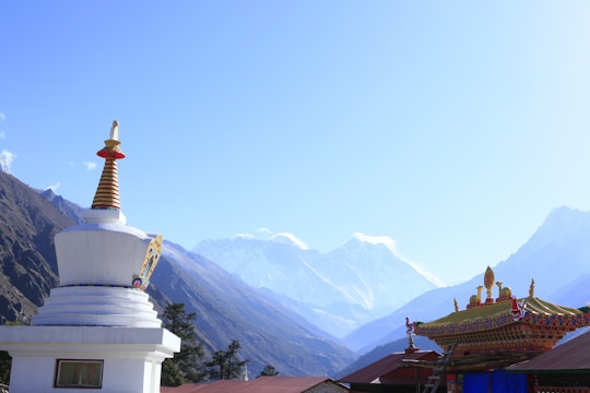 A stunning mountain landscape features snow-capped peaks in the background. In the foreground, a white Buddhist stupa with a golden spire and colorful decorations stands on the left, while a richly decorated temple roof is on the right. Trees can be seen at the base of the mountains.