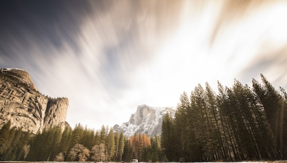 A scenic landscape featuring a valley flanked by tall, rocky mountains and dense pine forests. The sky above shows dynamic cloud movement, suggesting a long exposure photograph. The sun illuminates the scene, casting soft light on the rock formations and trees.