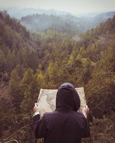 A rugged survivalist standing in a dense, misty forest wearing tactical gear and holding a map under a dark, stormy sky.