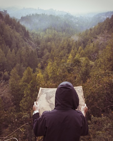 A person wearing a hooded jacket is holding a map, standing in front of a lush, green forest landscape. The scene is set in a mountainous area with a misty atmosphere, suggesting an exploration or adventure.