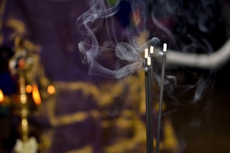 Four incense sticks releasing swirling smoke in front of a blurred background with warm lighting, possibly a brass lamp.