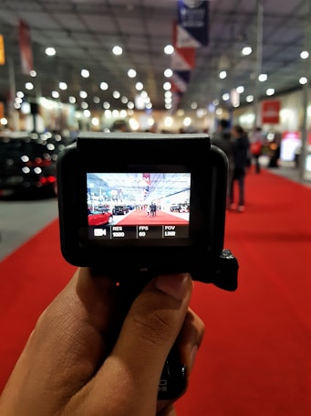 A hand holds a camera with a screen showing a scene at an indoor event. The event space has a red carpet and displays with people and bright lighting throughout the hall. The camera screen captures a perspective of the event, providing a clear internal view.