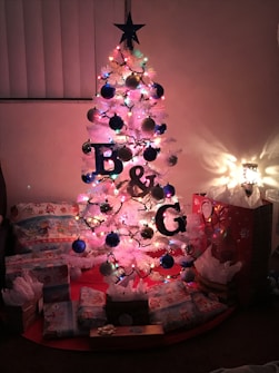 A small, white artificial Christmas tree adorned with multicolored lights and blue, gold, and red ornaments stands in a dimly lit room. The letters B & G are prominently displayed on the tree. Underneath, several wrapped presents in festive paper and gift bags are scattered around.