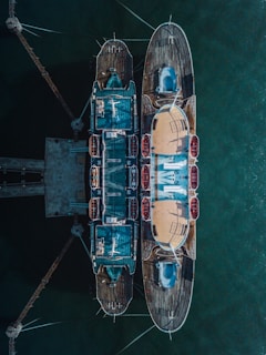 Aerial view of a docked, symmetrical nautical vessel. The ship is intricately designed with multiple decks and visible lifeboats on the sides. The calm greenish water surrounds the vessel, creating a serene atmosphere.