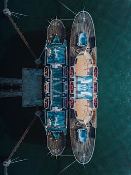 Aerial view of a docked, symmetrical nautical vessel. The ship is intricately designed with multiple decks and visible lifeboats on the sides. The calm greenish water surrounds the vessel, creating a serene atmosphere.