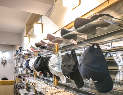 A retail display features a variety of caps organized on glass shelves, with colors including black, white, and pink. Additional accessories such as sunglasses and keychains are arranged below. The store has modern lighting and a dreamcatcher hanging on one side.