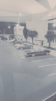 A parking lot with several vintage cars lined up beside a modern, minimalist building. Trees flank the building and the scene has a muted, retro appearance.