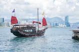 A traditional wooden junk boat with striking red sails is sailing in a body of water. In the background, there's a cityscape featuring modern skyscrapers and mountains under a partly cloudy sky. The boat, adorned with ornaments, adds an element of historical charm against the urban skyline.