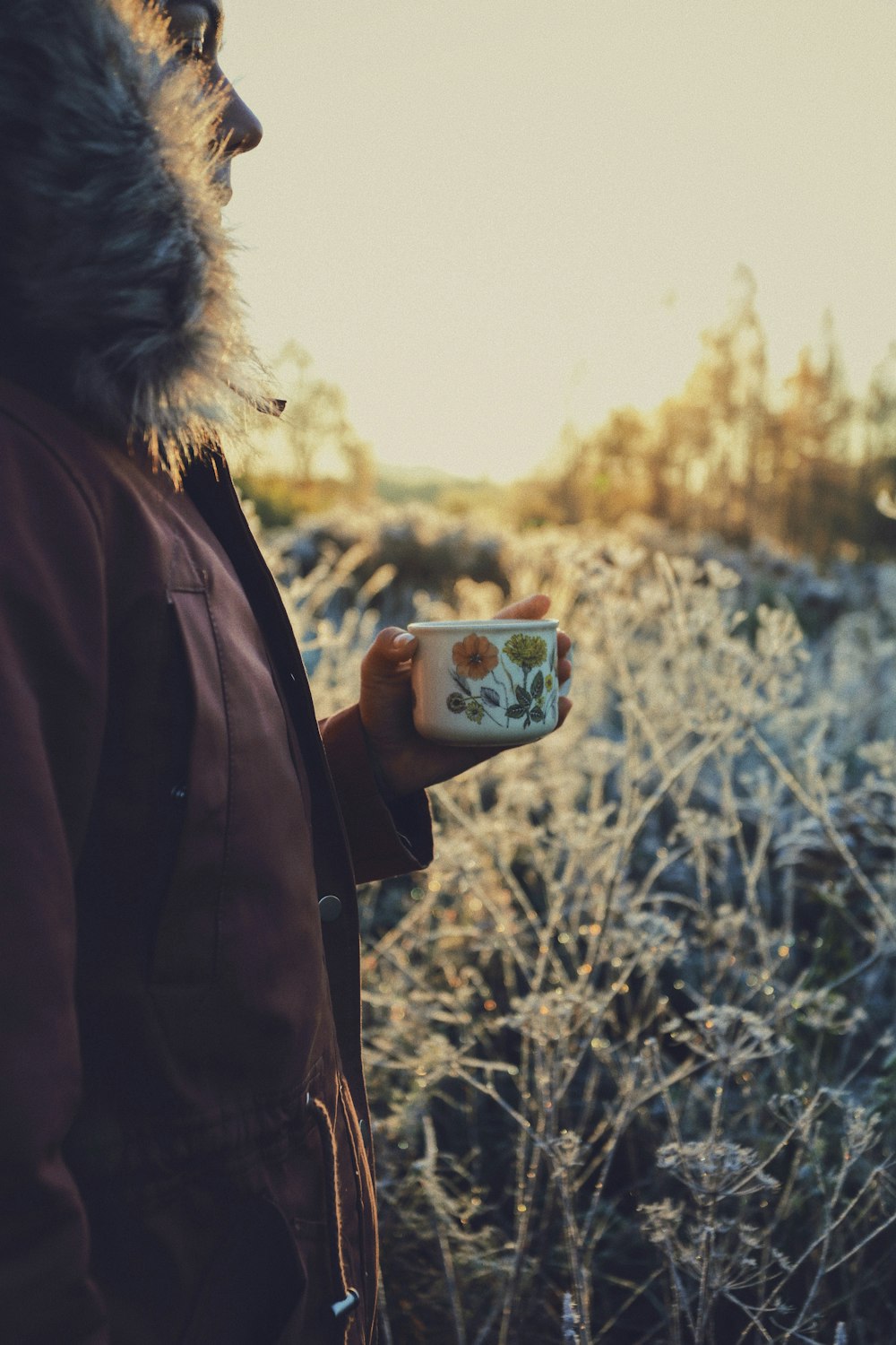 person in red fur-lined jacket holding white and green floral mug